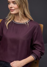Blusa de color púrpura oscuro con una textura suave, que presenta un escote redondeado y un dobladillo blanco en contraste. Mangas largas con puños ajustados.