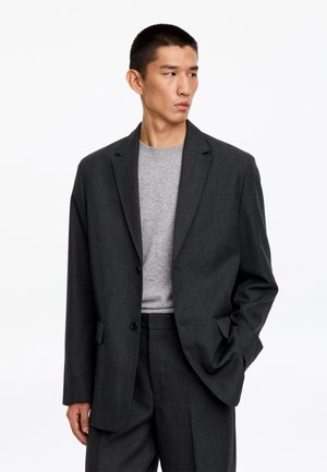 Homme portant un costume gris foncé avec un pull gris clair en dessous, debout avec une main dans la poche et regardant sur le côté.