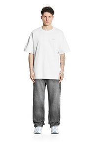 Biała bawełniana koszulka z luźnym krojem, zestawiona ze szarymi dżinsami. Posiada prosty design i widoczne tatuaże na przedramionach.