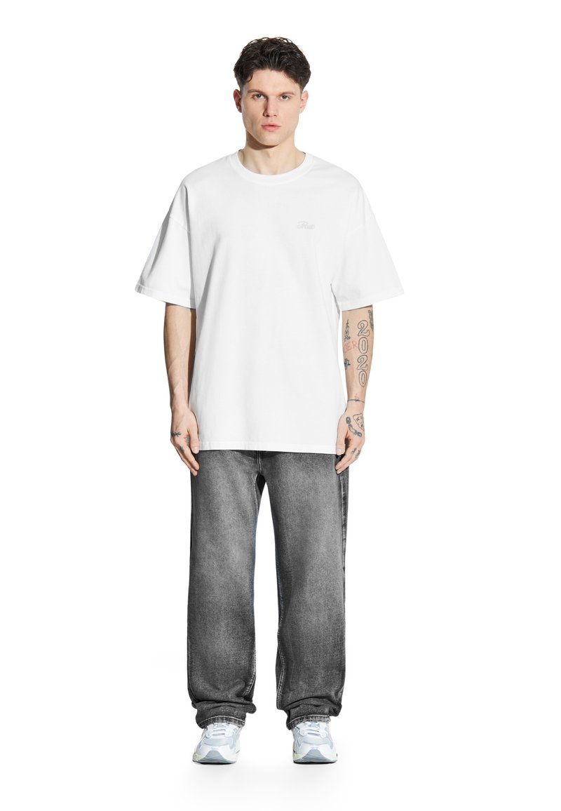 Biała bawełniana koszulka z luźnym krojem, zestawiona ze szarymi dżinsami. Posiada prosty design i widoczne tatuaże na przedramionach.
