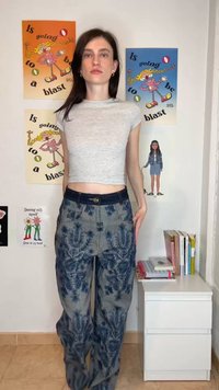 Frau mit langen dunklen Haaren, trägt ein graues, kurzes T-Shirt und blaue, mit Blumen gemusterte Hosen, steht vor einer Wand mit bunten Postern und Büchern auf einem weißen Nachttisch.