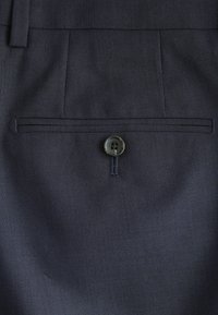 Gros plan sur un tissu de costume bleu marine foncé montrant une poche arrière avec un seul bouton noir et un détail de passants de ceinture au-dessus.