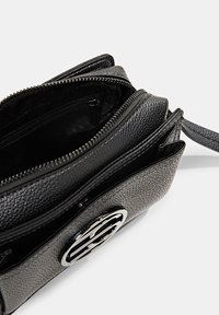 Černá texturovaná kožená taška přes rameno s několika zipovými kapsami a stříbrným logem na přední straně. Uvnitř je podšita černou tkaninou.