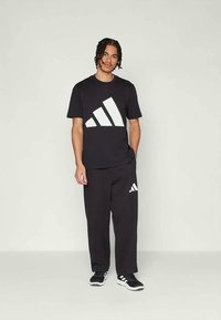 Svart atletisk outfit bestående av en kortärmad t-shirt med en stor vit adidas-logo och matchande byxor med logotypdetaljer.