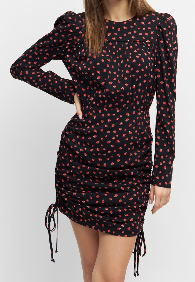 Femme portant une mini-robe noire à manches longues avec imprimé floral rouge et côtés froncés attachés avec de petits nœuds.