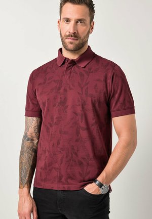 Mann mit kurzem Haar und Bart, der ein burgunderfarbenes, gemustertes Polohemd, schwarze Hosen, eine Armbanduhr trägt und ein Tattoo am linken Arm hat.