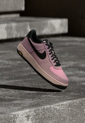 Różowo-czarny but sportowy Nike Air z czarnym logo i beżową podeszwą, zawieszony nad betonowymi schodami o szarej, teksturowanej powierzchni.