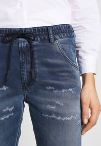 Henkilö, jolla on päällään siniset kulutetut farkut, joissa on elastinen vyötärö ja musta nauha, sekä valkoinen pitkähihaisessa paidassa, käsi lepää kyljessä.