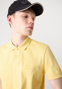 Gul polo skjorte med teksturert stoff, to knapper og en liten preget logo på venstre bryst. Svart caps med kontrasterende hvite detaljer.