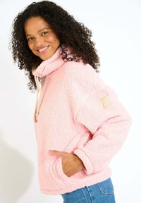 Pullover in pile rosa con colletto alto, tasche laterali e una texture morbida. Presenta una patch con logo sulla manica. Vestibilità casual e ampia.