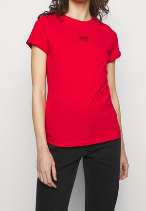 Kobieta w czerwonym T-shircie z krótkim rękawem z podwiniętym mankietem i naszywką z logo "HUGO" na piersi, w połączeniu z czarnymi spodniami.