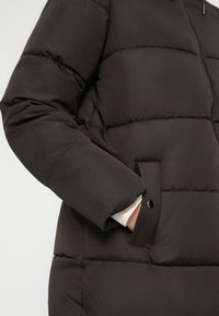 Giubbotto a vento marrone con motivo a quadrati orizzontali, cappuccio removibile, tasche laterali e finitura liscia. Presenta una chiusura con zip e bottoni a scatto.