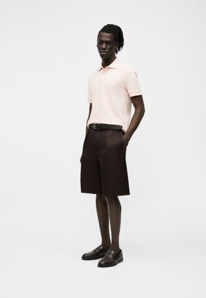 Man met dreadlocks die een lichtroze polo, donkerbruine knielange shorts, een zwarte riem en zwarte loafers draagt, staat met zijn handen in zijn zakken.