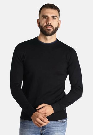 Uomo barbuto con capelli corti che indossa un maglione nero a maniche lunghe con scollo rotondo e jeans blu chiaro, in piedi con le mani giunte.