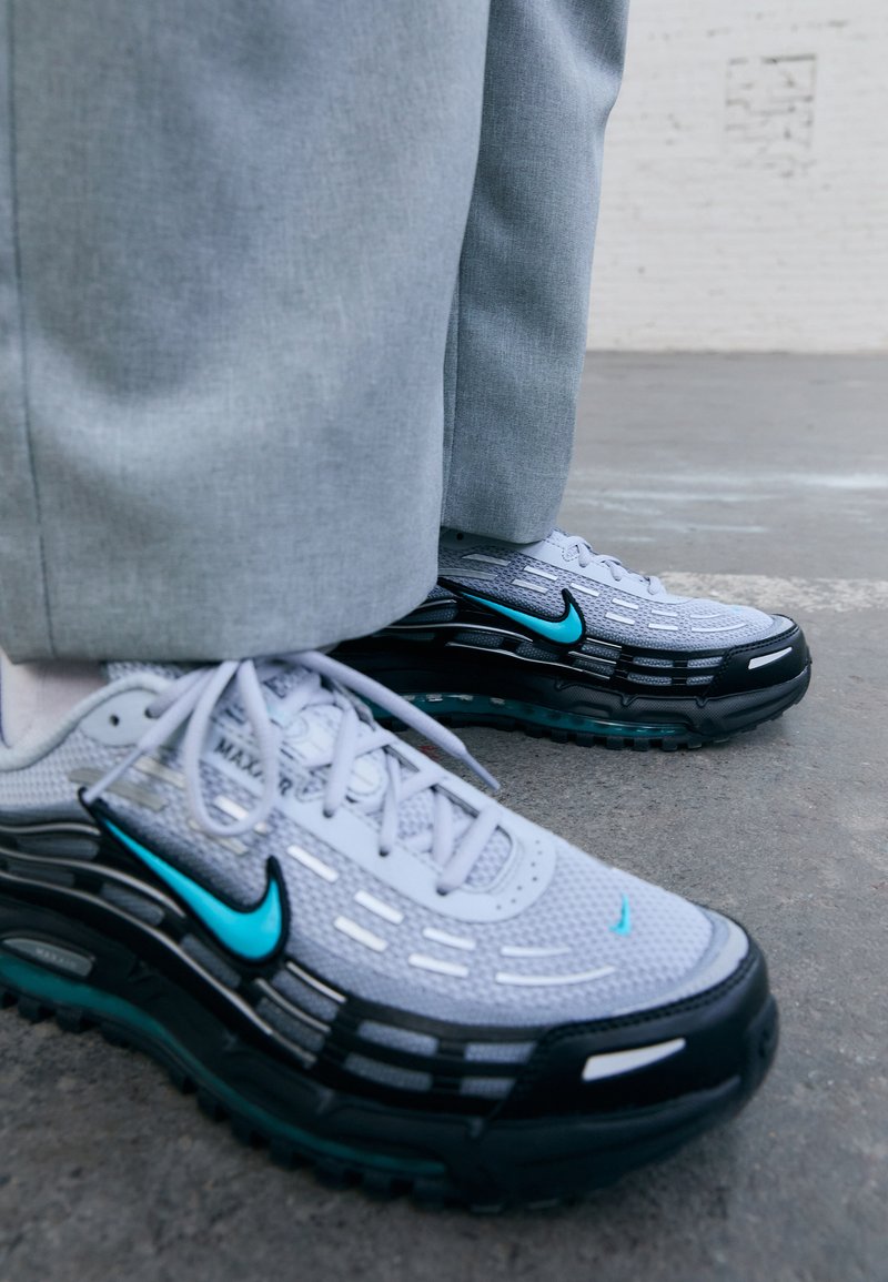 Nike sporta apavi ar pelēku acs augšu, melnu gumijas zoli ar tirkīza akcentiem un atstarotāju detaļām. Saiņojuma dizains ar redzamām zīmola atzīmēm.