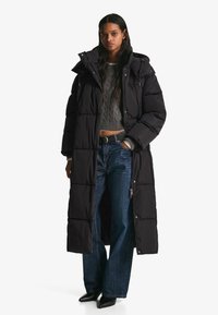 Geacă lungă neagră matlasată, cu glugă și model în carouri, asortată cu un pulover gri cu torsadă și blugi albaștri cu picioare largi. Pantofi negri cu vârf ascuțit.