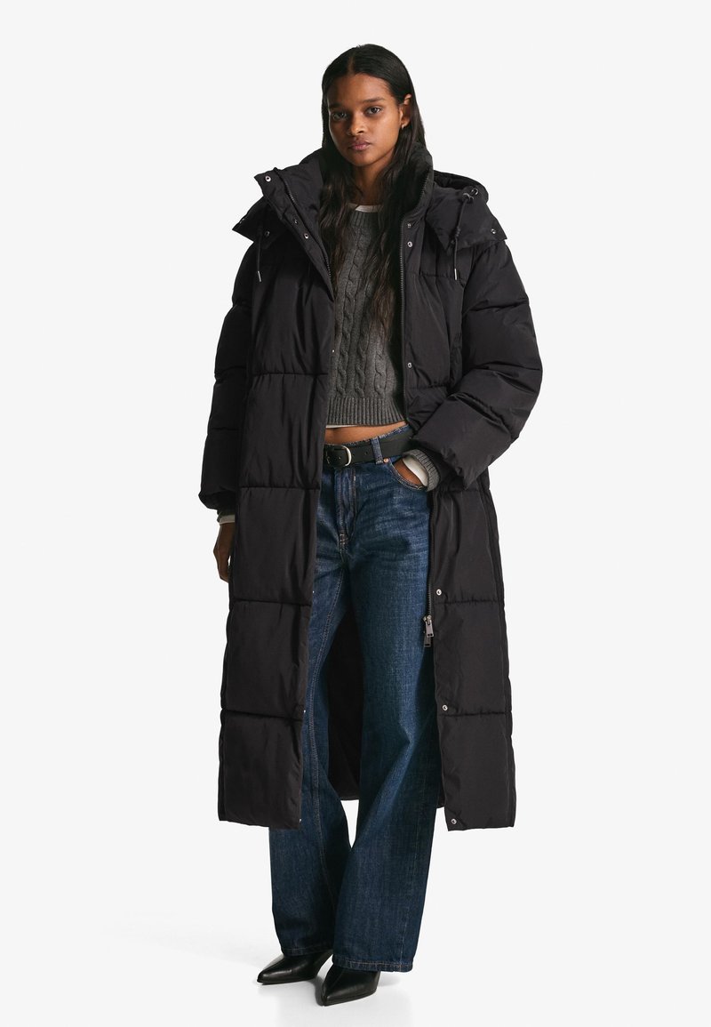 Geacă lungă neagră matlasată, cu glugă și model în carouri, asortată cu un pulover gri cu torsadă și blugi albaștri cu picioare largi. Pantofi negri cu vârf ascuțit.