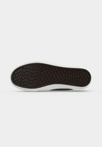 Semelle de chaussure avec motif gaufré noir et bord blanc sur un fond clair uni.