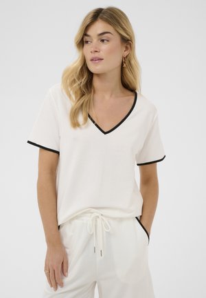 Femme blonde portant un haut blanc à col en V et manches courtes ainsi qu'un pantalon assorti avec un bord noir, regardant sur le côté, mains dans les poches.