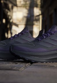 Par af sorte og lilla Adidas løbesko med "Light Boost" sål på en brostensbelagt gade i blødt naturligt lys.