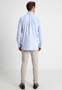 Camisa de algodón azul claro de manga larga con cuello abotonado y pliegue en la espalda, combinada con pantalones beige y zapatos negros.
