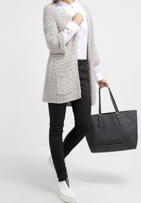 Ljusgrå stickad cardigan med öppen front och ficka, ihop med svarta jeans och vita slip-on skor, håller en svart tote-väska.