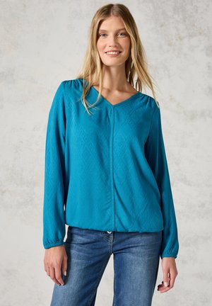 Blusa lunga teal con scollatura a V, motivo a rombi testurizzato, maniche elastiche e cucitura centrale. Abbinata a jeans blu.