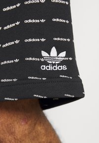 Svarta shorts med ett upprepande mönster av vit Adidas-logotyp, tillverkade av mjukt tyg med en broderad logotyp på fållen.