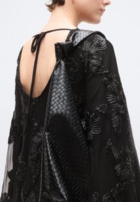 Μαύρο φόρεμα με κεντήματα λουλουδιών, ανοιχτή πλάτη και υφασμένη, υφασμένη διακόσμηση στον ώμο. Ρυθμιζόμενο κορδόνι στον λαιμό για επιπλέον λεπτομέρεια.