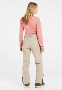 Lichtgewicht, lange mouw, roze top gecombineerd met beige skibroek met een gestructureerde tailleband en ritssluiting bij de enkels, gedragen met groene laarzen.