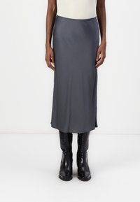 Osoba w szarej midi spódnicy i czarnych skórzanych butach o fakturze, stojąca na tle białego tła.