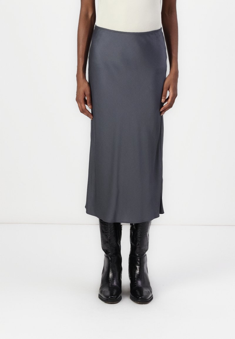 Osoba w szarej midi spódnicy i czarnych skórzanych butach o fakturze, stojąca na tle białego tła.