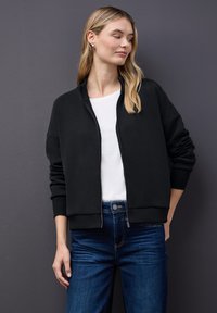 Chaqueta bomber negra con cremallera en el frente, puños acanalados y una textura suave, combinada con una camiseta blanca y jeans azules.
