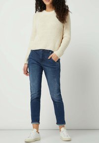 Cream knitted sweater with a textured pattern, paired with dark blue slim-fit denim jeans. White low-top sneakers complete the outfit.