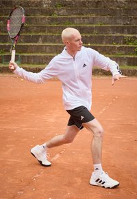 Tenisový hráč v bílé bundě adidas a černých šortkách, držící raketu, na červeném antukovém kurtu s kroky v pozadí.