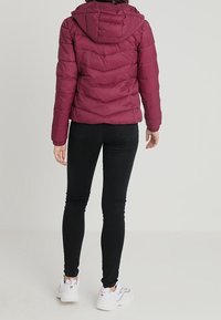 Maroon puffer jacket with a hood, featuring a quilted design and elastic cuffs, worn over black skinny jeans and white sneakers.