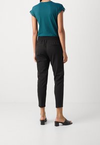 Donna con indosso una maglietta teal a maniche corte, pantaloni neri a vita alta e tapered, e sandali con tacco aperti sul retro neri, in piedi contro uno sfondo bianco semplice.