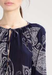 Marineblaue Bluse mit aufwendigem weißen Paisleymuster, leichtem Stoff, runder Halslinie und Bindeschlaufe vorne.