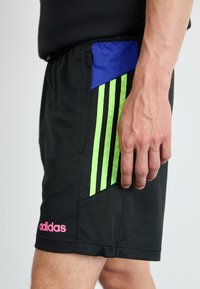 Melni sporta īsie šorti ar teksturētu audumu, zaļām vertikālām svītrām un zilu kabatas akcentu. Rozā Adidas logo malā.