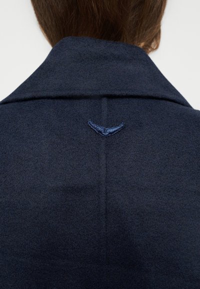 Σκούρο ναυτικό μπουφάν πίσω με μικρή ραμμένη λεπτομέρεια σε σχήμα φτερού κάτω από το γιακά σε άτομο με ανοιχτά καστανά μαλλιά.