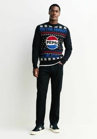 Schwarzer Strickpullover mit festlichem Design, das das Pepsi-Logo in Rot, Weiß und Blau zeigt, kombiniert mit dunklen Hosen und schwarzen Sneakern.
