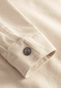 Close-up af en beige stofmanchet med en sort trykknap indgraveret med "G-ABSA" på et langærmet tøjstykke.