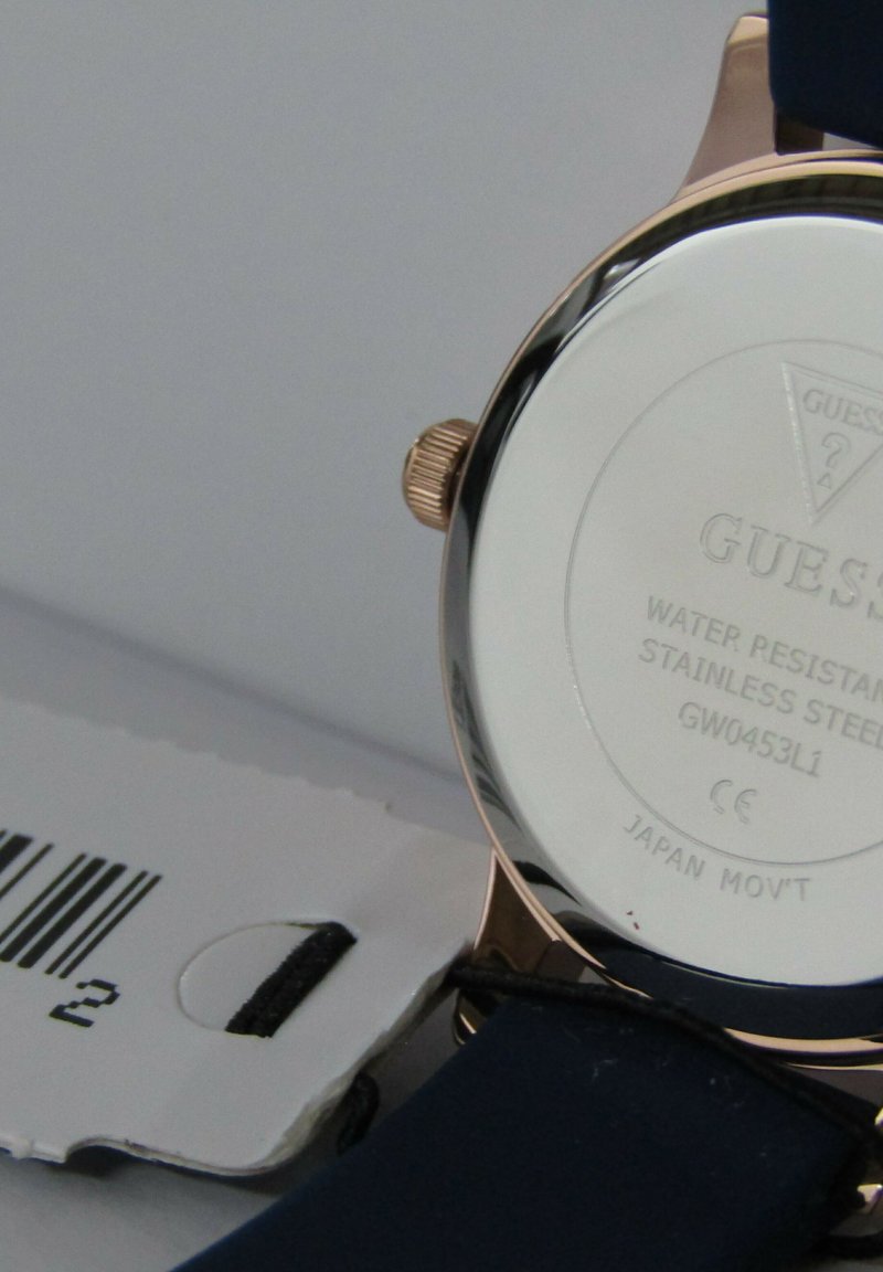 Reloj Guess con fondo de acero inoxidable, acentos en oro rosa y correa azul marino. Presenta resistencia al agua y una etiqueta de código de barras marcada.