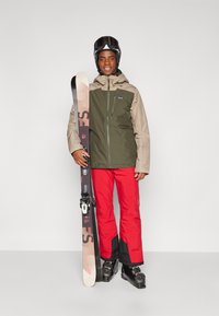 L'équipement de ski comprend une veste olive et beige, un pantalon rouge vif, des lunettes de ski et des skis avec des fixations noires et un motif géométrique.