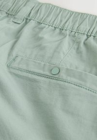 Tissu vert clair d'un pantalon à taille élastique montrant une poche arrière cousue avec un rabat sécurisé par un bouton-pression assorti.