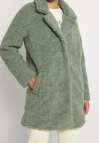 Grön, fluffig kappa i strukturerat material, med krage, öppen front och sidofickor. Lämplig för avslappnad användning i svalt väder.