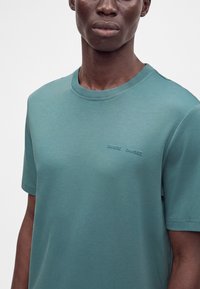Homme portant un t-shirt uni turquoise à manches courtes avec un petit texte en relief sur le côté gauche de la poitrine sur fond blanc.