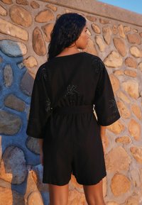 Cat suit nero con maniche corte e motivi di palme ricamati. Presenta una vita elastica e una vestibilità comoda, realizzato in un tessuto leggero.