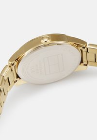 Orologio in acciaio inossidabile dorato con cassa rotonda, lunetta liscia e bracciale a maglia. Presenta una corona texturizzata e un fondello trasparente.