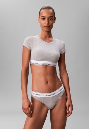 Vrouw draagt een grijs ingekort T-shirt van Calvin Klein en bijpassend ondergoed met witte merkemblemen op de tailleband, staand tegen een effen grijze achtergrond.